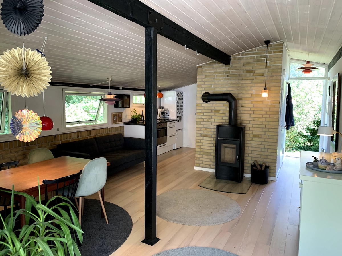 An open living space is highlighted by a modern design featuring a large sectional sofa and a dining table with mixed seating. Natural light floods the area through large windows, while a cozy wood-burning stove is integrated into the brick wall, and plants add a touch of greenery.