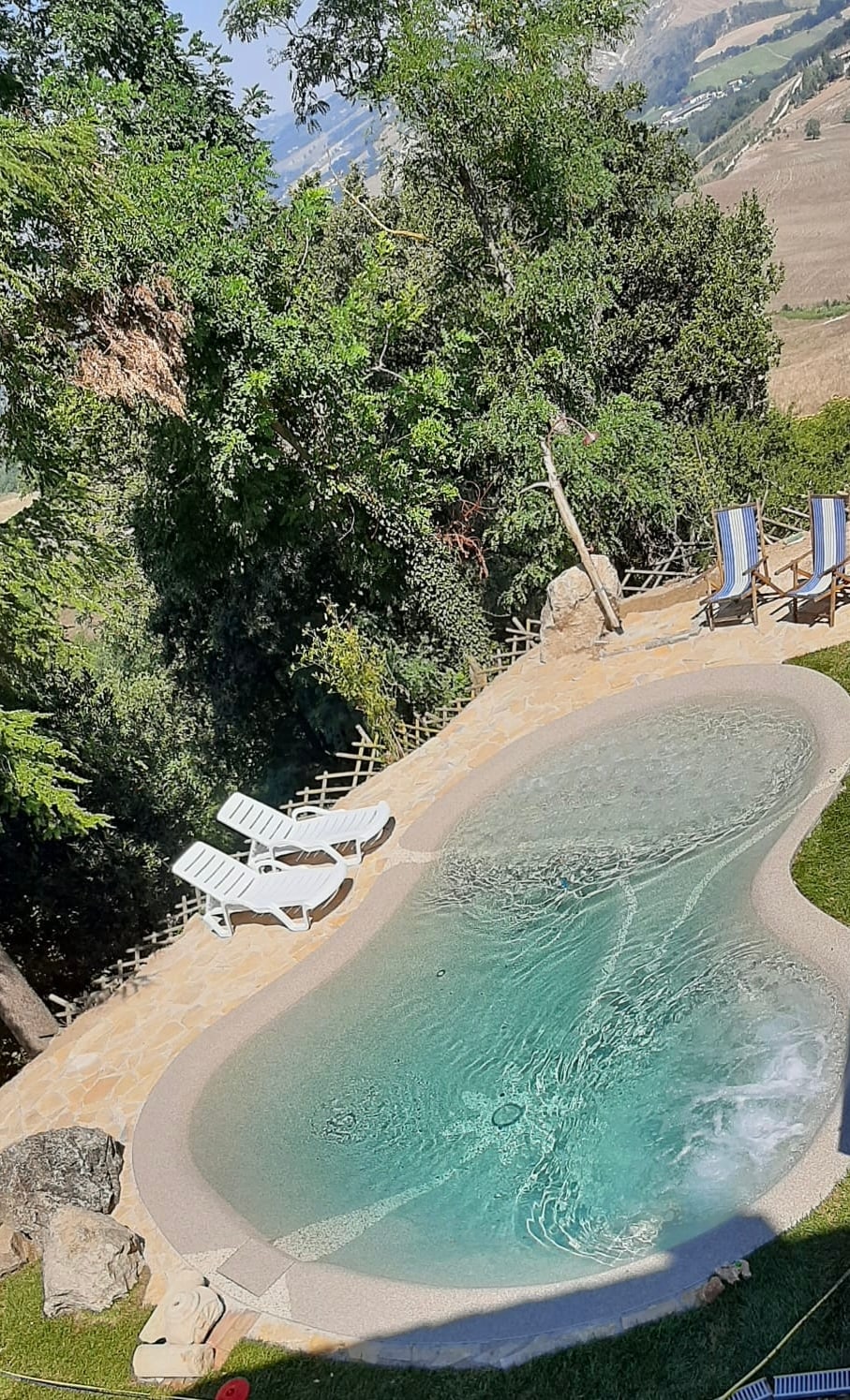 A small, inviting biolake features clear water and is surrounded by lush greenery. Two white lounge chairs are positioned nearby, offering a serene spot for relaxation. The natural stone edges blend harmoniously with the landscape, creating a tranquil outdoor atmosphere.