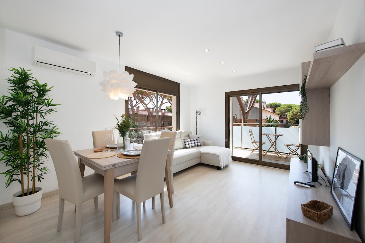 A spacious living and dining area is highlighted by natural light from large glass doors leading to a terrace. A dining table with four chairs is positioned alongside a comfortable seating area. Decorative plants add a touch of greenery to the serene environment.