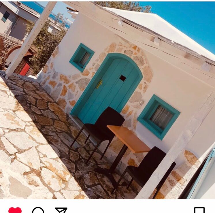 A quaint exterior view features a stone pathway leading to a charming entrance, adorned with a blue door. Two dark chairs and a small table are placed on a covered patio area, inviting outdoor relaxation. The surrounding white walls enhance the bright and airy atmosphere.