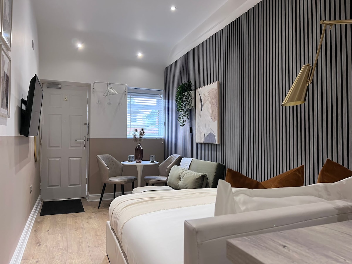 A modern studio apartment features minimalist decor with neutral tones. A spacious bed is positioned near a sleek wall, complemented by elegant accent pillows. A small dining table and two chairs are set by a window, while a functional entrance is visible at the far end.