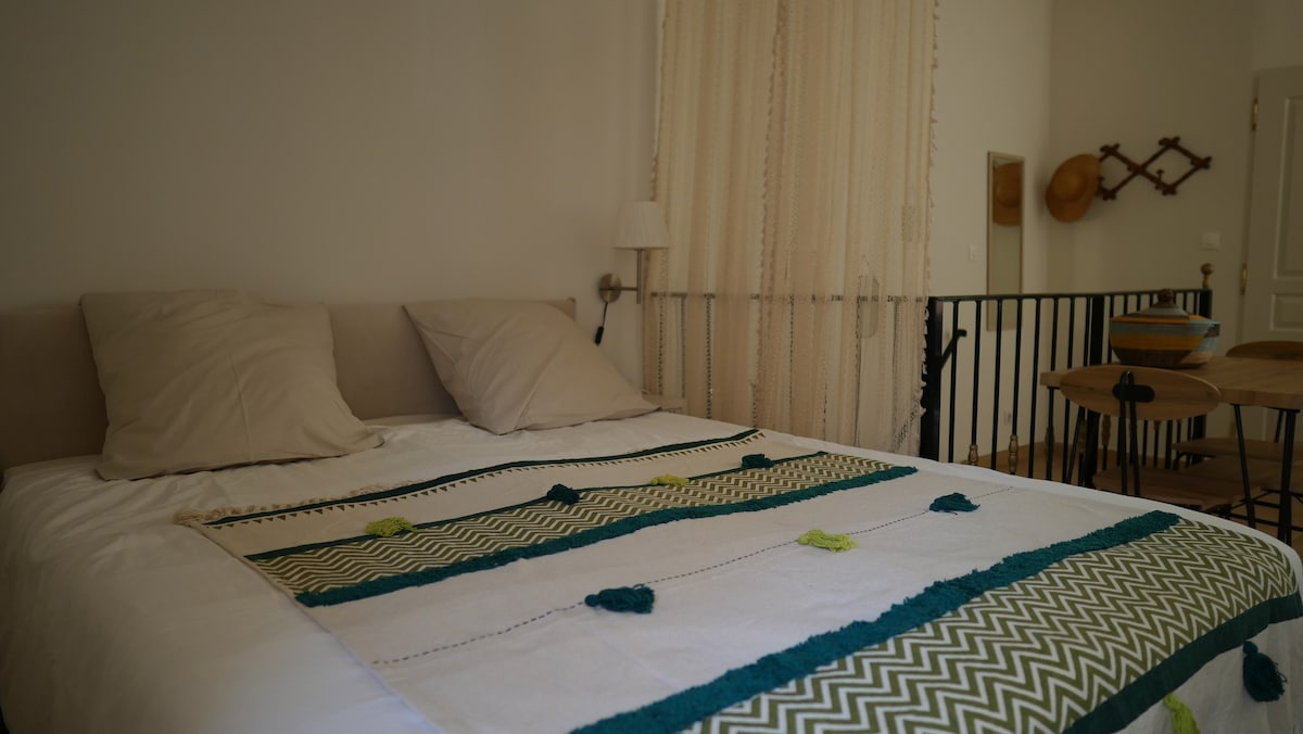 A cozy sleeping area features a spacious bed adorned with a patterned blanket in green and white. Two soft pillows rest at the headboard. A lightweight curtain filters natural light, while simple decor is seen in the background.