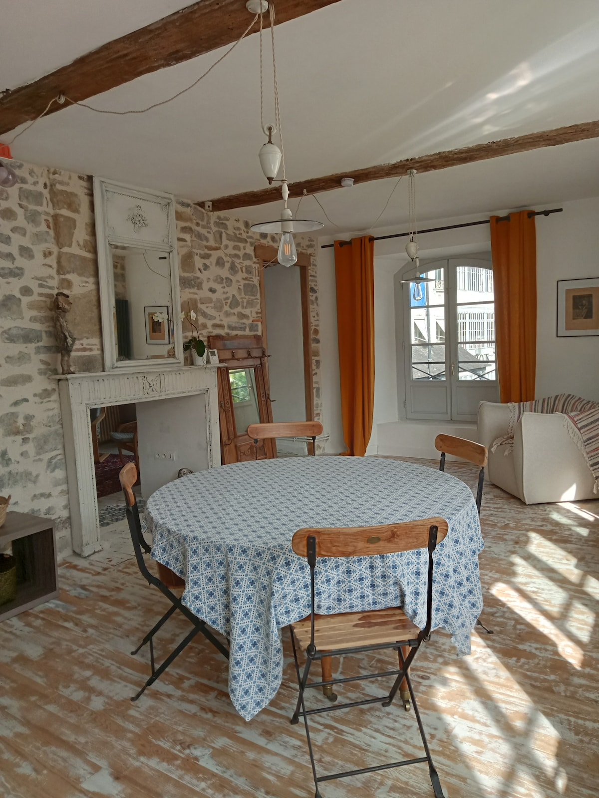 A round dining table is set with a patterned tablecloth, surrounded by wooden chairs. Exposed stone walls and wooden beams add character to the space. Natural light filters in through a window adorned with orange curtains, highlighting a cozy seating area nearby.
