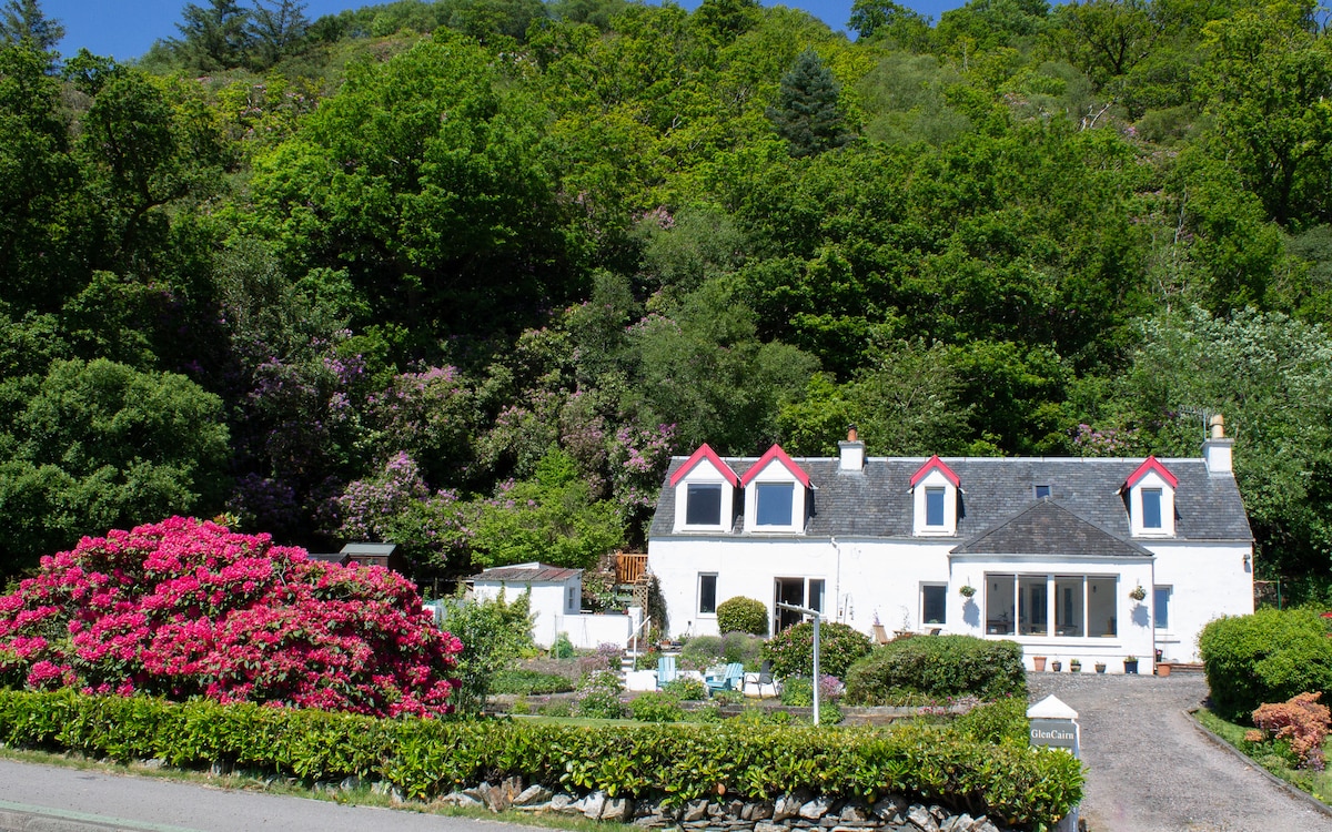 The exterior of Glencairn Flat features a charming white structure with red accents and a landscaped garden. Lush greenery surrounds the property, including vibrant flowering shrubs. A gravel driveway leads to the entrance, providing a welcoming approach to the home.