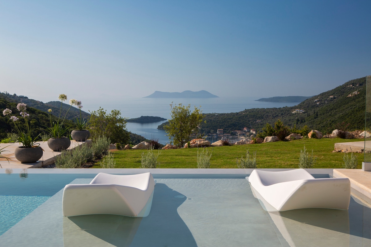 Two modern lounge chairs are positioned in a tranquil swimming pool, overlooking a serene landscape. Expansive views of the sea and distant islands can be seen in the background, complemented by lush greenery and carefully arranged potted plants along the edge of the property.