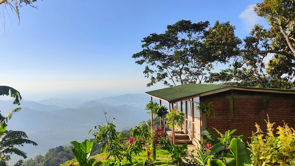 A private bungalow is positioned on a hillside, surrounded by lush tropical foliage. Large windows offer an unobstructed view of the mountains and sky, showcasing the vast landscape below. The green roof complements the natural surroundings, enhancing the serene atmosphere.