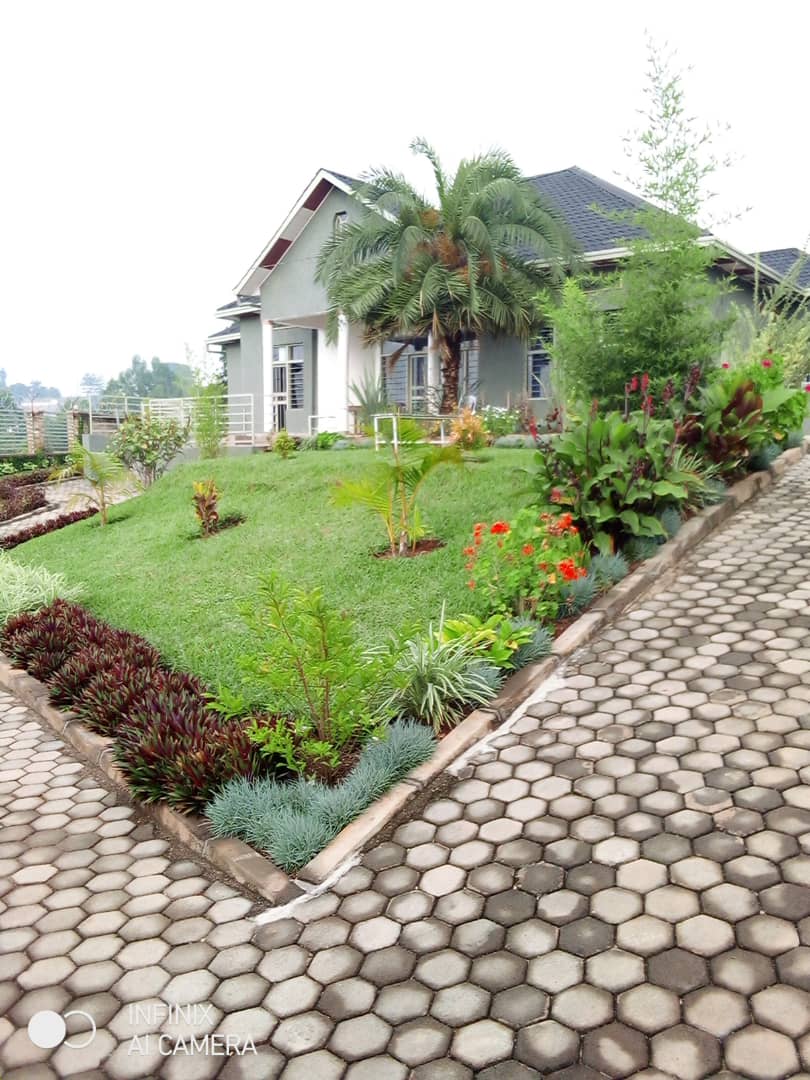 A well-maintained exterior showcases a charming house surrounded by lush greenery. Colorful flowers and neatly trimmed shrubs enhance the landscape, while a patterned stone driveway leads to the entrance. A backdrop of trees complements the peaceful setting.