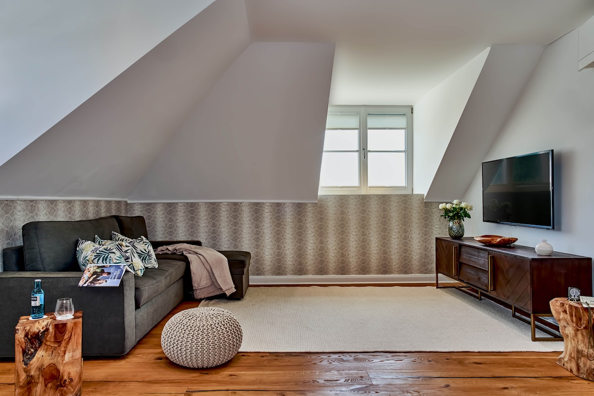 An airy living area features a modern gray sofa paired with decorative pillows. A plush pouf is positioned on a light area rug, and two wooden side tables are set with drinks. A television is mounted on the wall opposite a large window, allowing ample natural light.