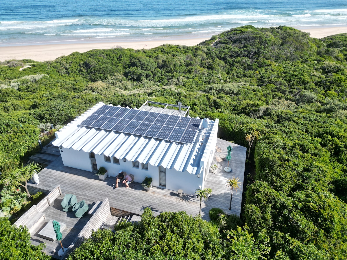 An aerial view of a white cottage surrounded by lush greenery and fynbos, showcasing a wooden deck with seating areas. Solar panels are visible on the roof, indicating sustainable energy use. The coastline and beach stretch out in the background, adding to the serene landscape.