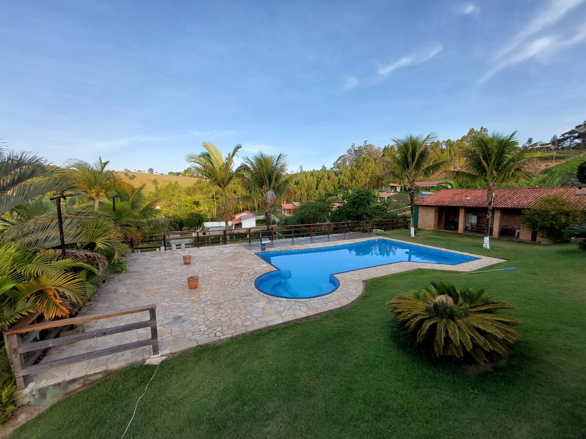 A large swimming pool is set amid lush greenery and palm trees, surrounded by a stone patio. The inviting water reflects the clear sky, while a well-maintained lawn complements the tranquil outdoor setting. Rustic buildings can be seen in the background.