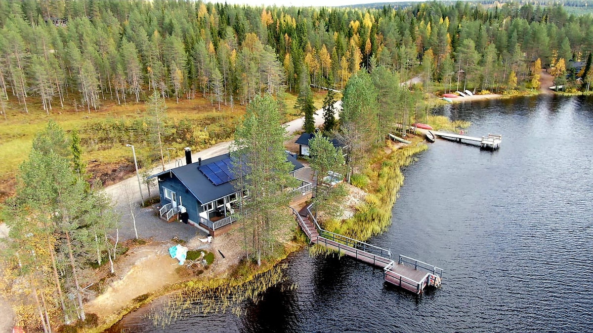 An aerial view captures a modern cabin by a serene lake, surrounded by lush greenery. A private dock extends into the water, with additional boats visible nearby. Solar panels are noted on the cabin's roof, contributing to a sustainable environment.