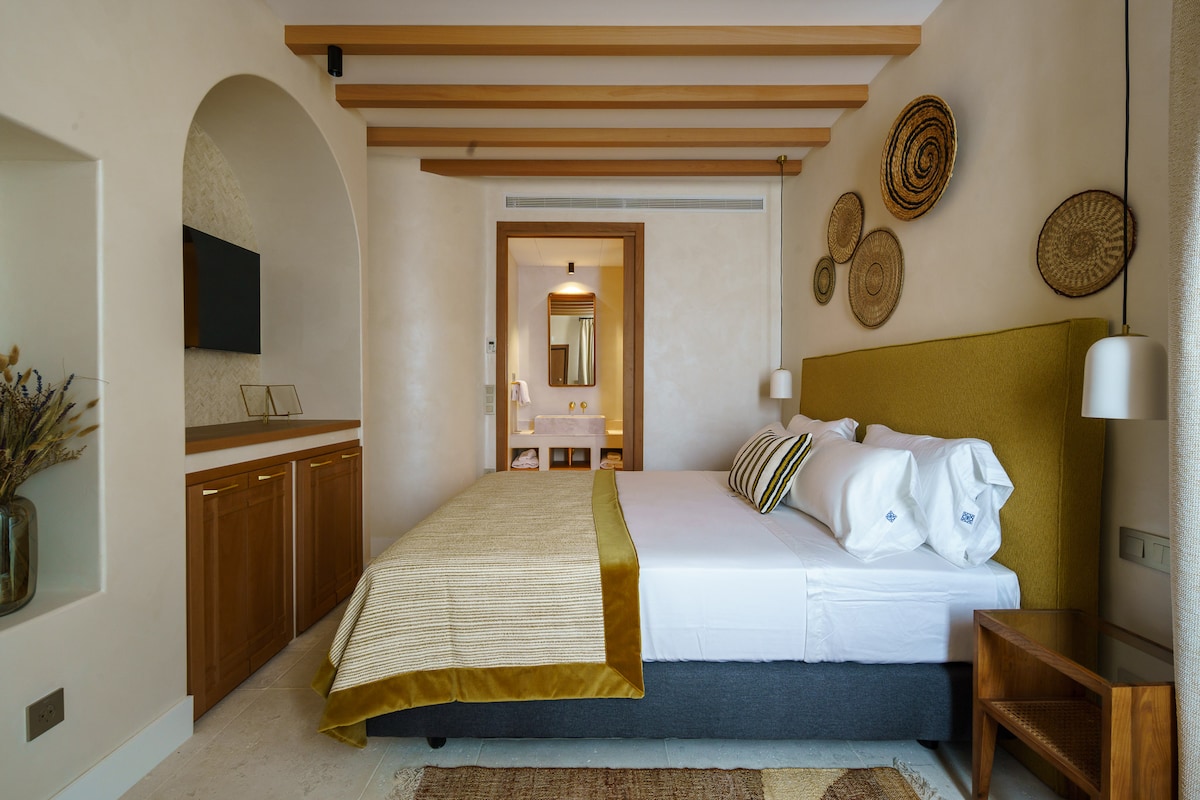A spacious sleeping area features a comfortable Queen Size bed with white linens and decorative throw pillows. Natural light enters through a nearby window. Woven wall art accents the warm-toned walls, while wooden beams and cabinetry add to the inviting design.