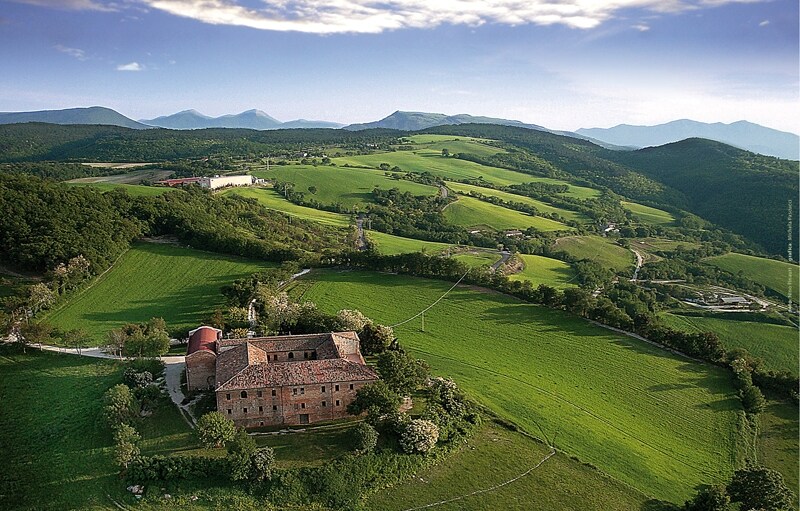 An expansive view of rolling green hills stretches across the landscape, showcasing the natural beauty of the region. A rustic building with a tiled roof is nestled among trees, while distant mountains form a serene backdrop under a partially cloudy sky.