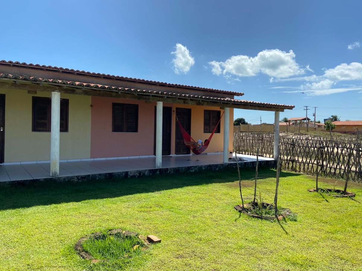 A single-story building is shown with a covered porch. A red hammock hangs between two posts, inviting relaxation. The lush green lawn features circular patches of grass, and a woven fence is visible in the background under a clear blue sky.