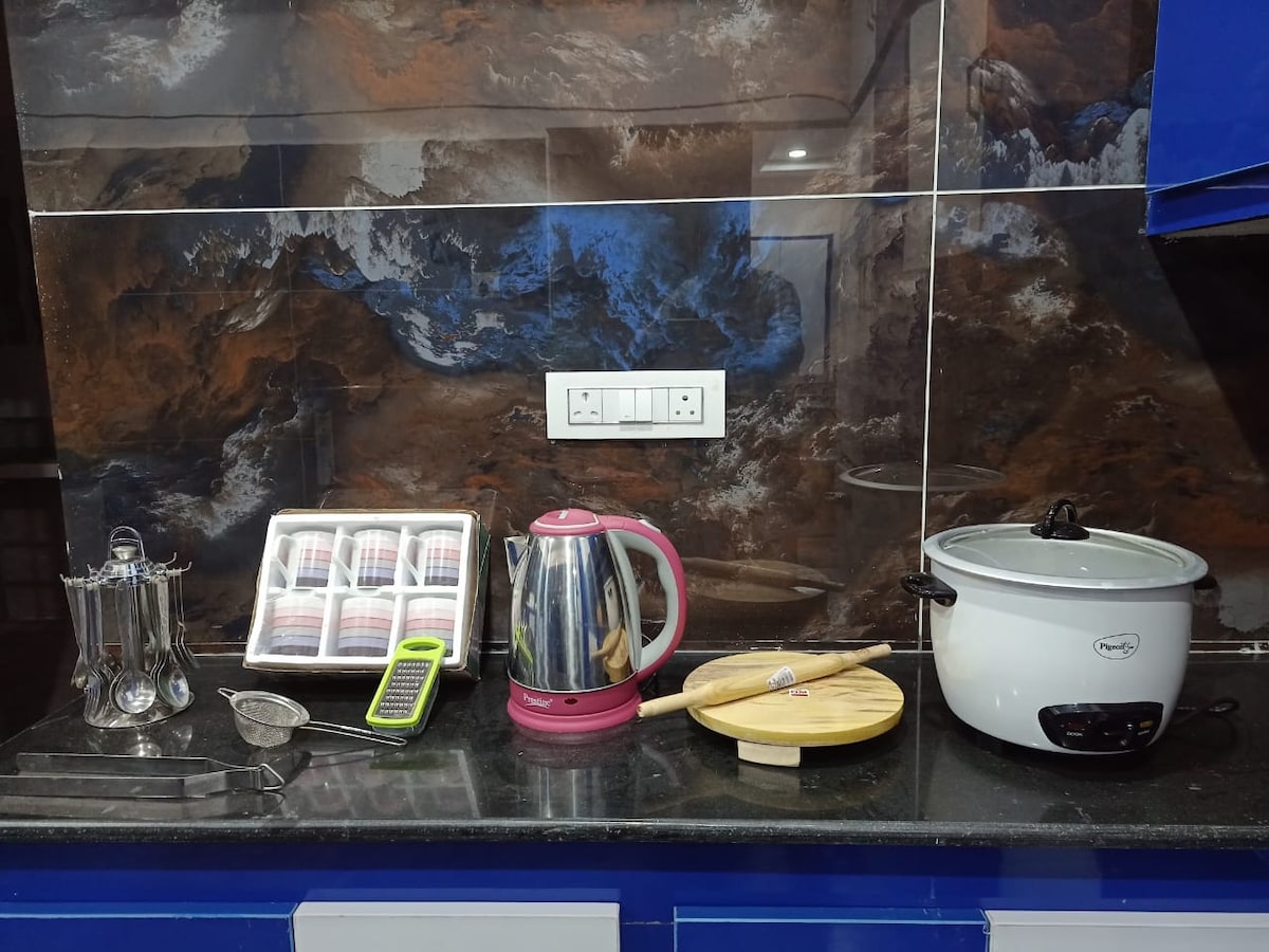 A modern kitchen surface is displayed featuring a stainless steel kettle, a slow cooker, and a wooden chopping board. A tea strainer and a green handheld scale are also visible, alongside a neatly arranged set of spice jars, creating an organized cooking space.