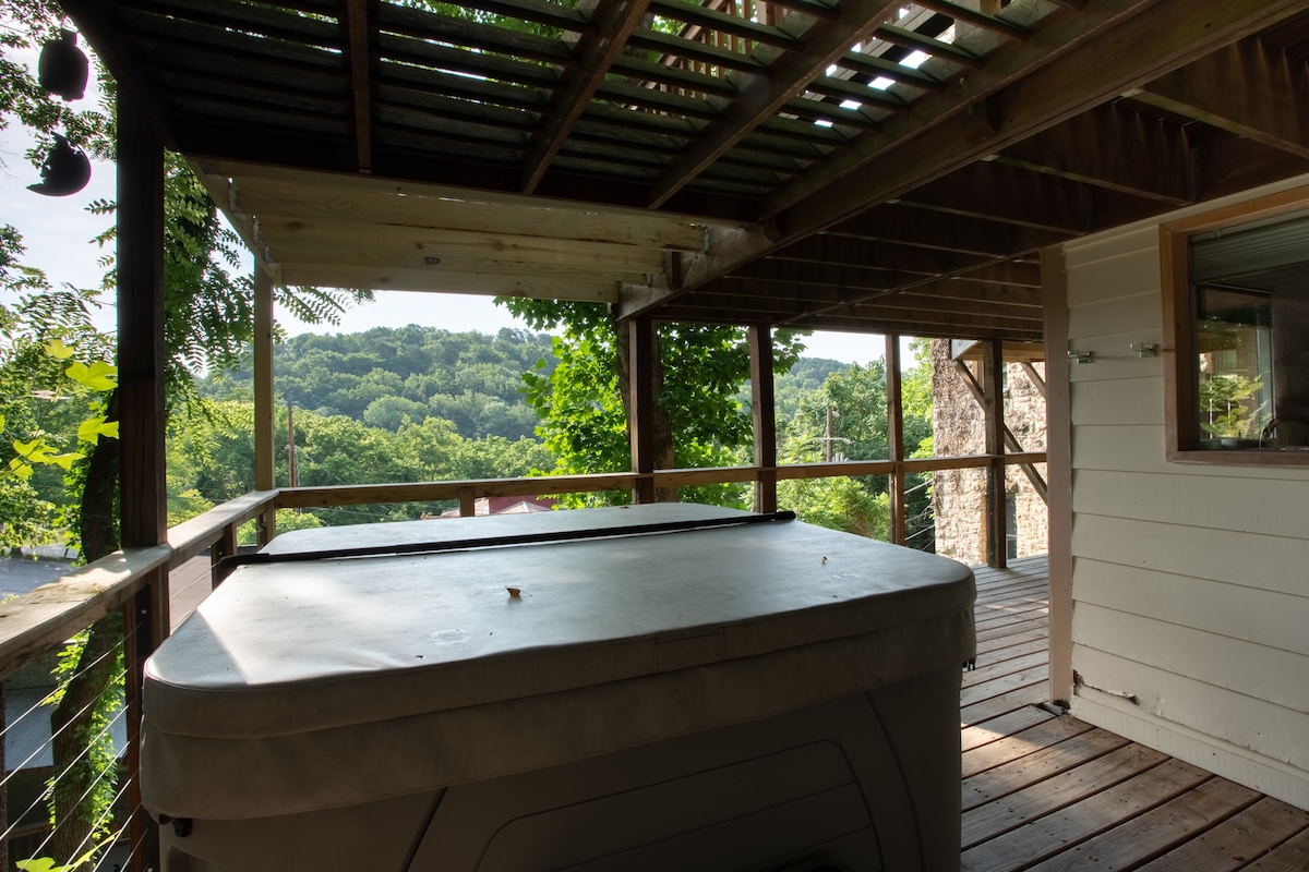 A spacious deck offers a hot tub, surrounded by wooden beams and a view of the lush landscape. Sunlight filters through the greenery, creating a serene atmosphere. The area is partially covered, providing a comfortable space for relaxation.