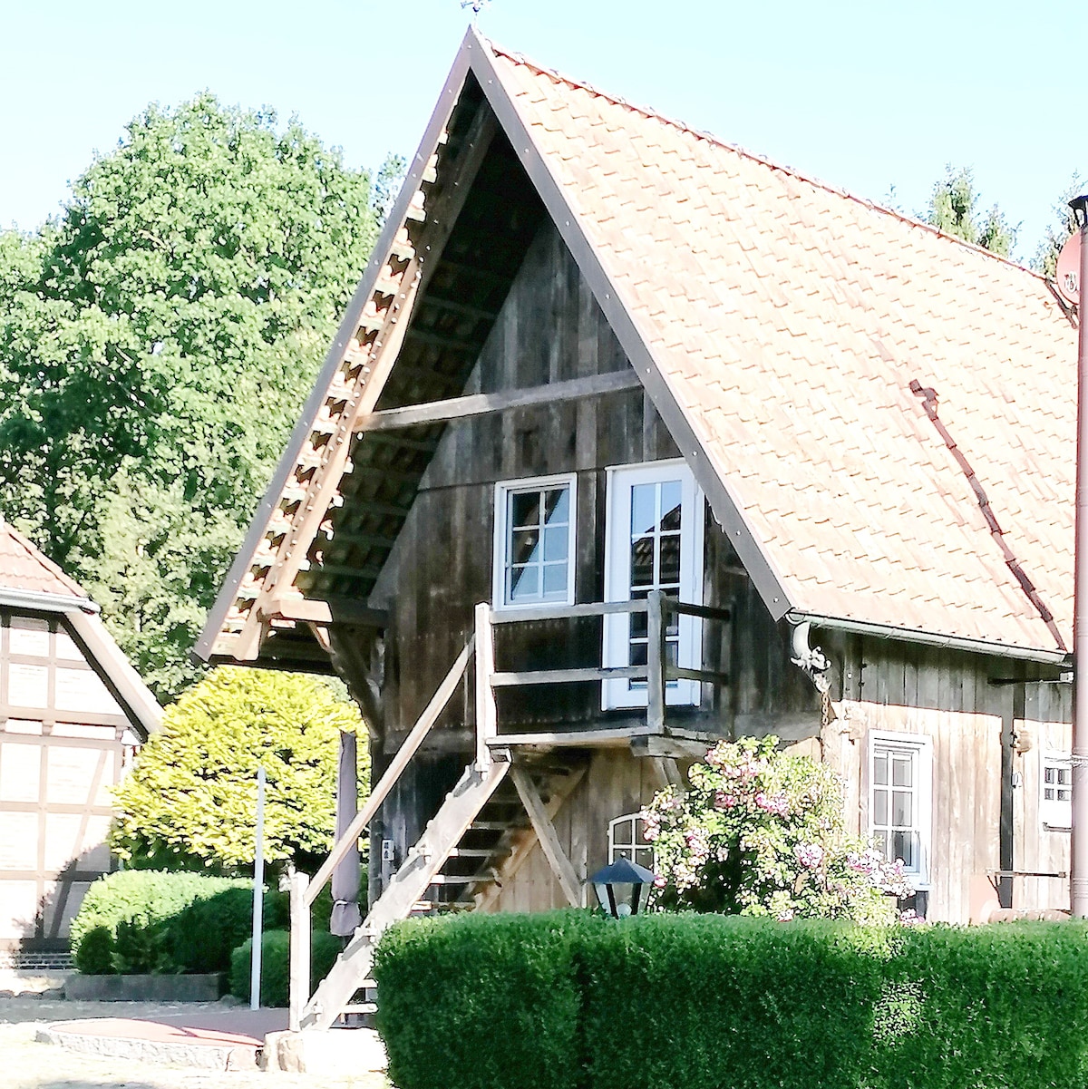 The exterior of the historic stair storage building is shown, featuring wooden beams and a steeply pitched roof. A staircase leads to a balcony, surrounded by manicured greenery and blooming flowers, creating a harmonizing appearance with the natural surroundings.