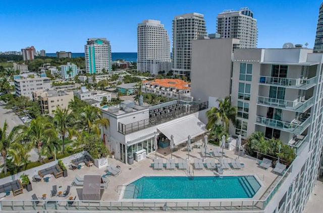 A scenic view of the rooftop pool area is presented, surrounded by lounge chairs and palm trees. The sparkling blue water of the pool invites relaxation, and the distant ocean can be glimpsed amid the coastal skyline, featuring various buildings under a clear blue sky.