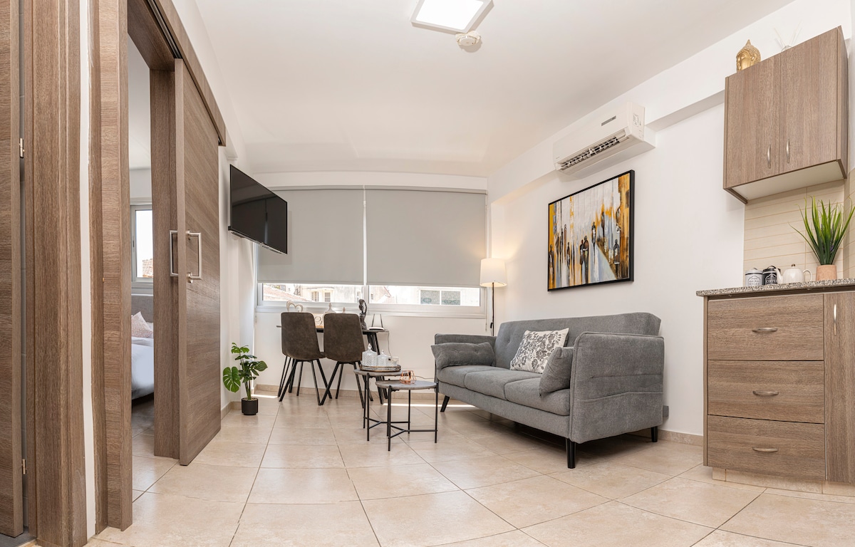 A modern living area features a grey sofa, accompanied by a coffee table. Two chairs and a small dining table are positioned nearby. A wall-mounted television and artwork add character to the space, while natural light filters through a window with light shades.