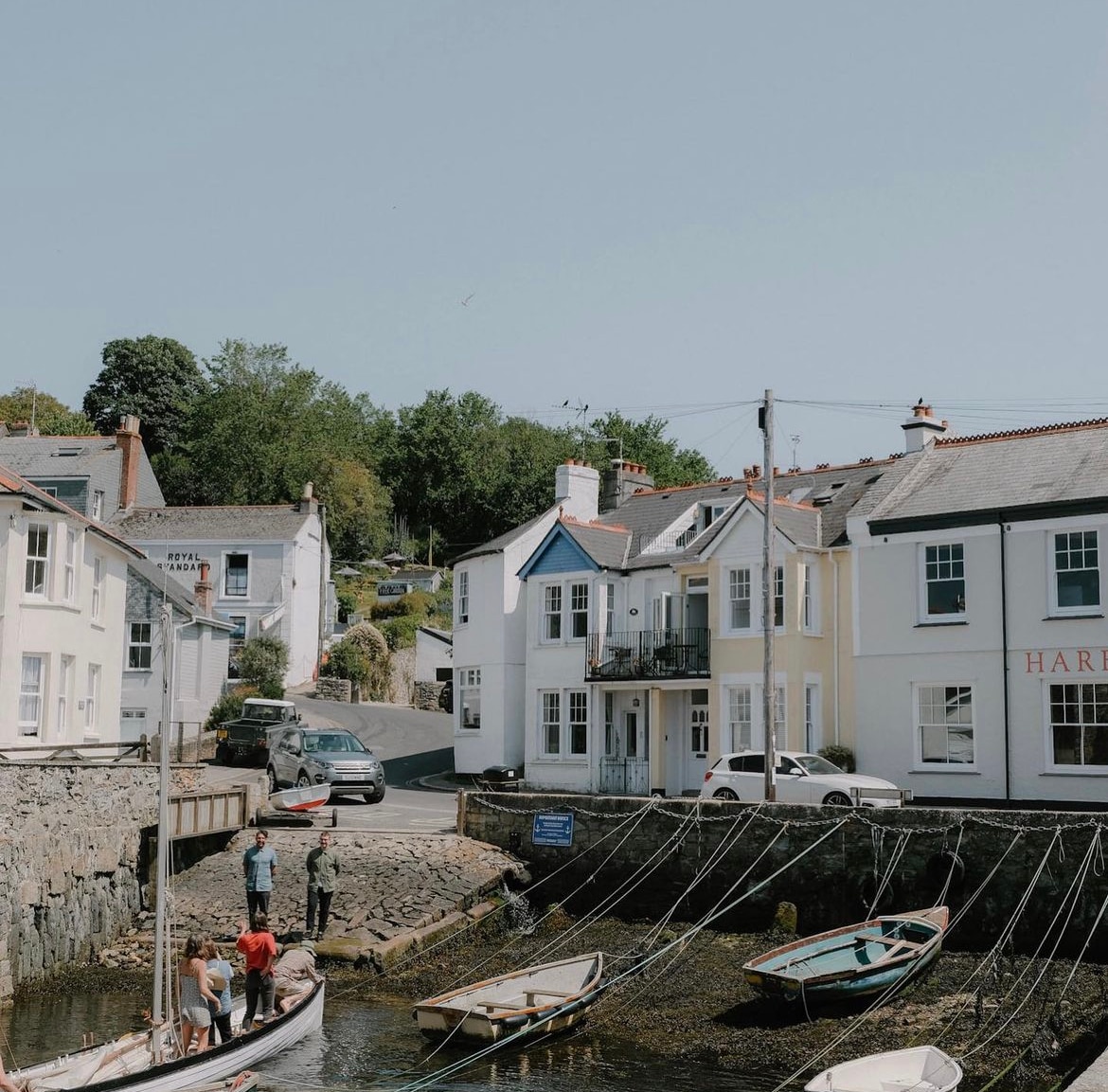 A scenic view of a tranquil quay, featuring white and pastel-colored houses lining the waterfront. Small boats are tied to wooden posts in the foreground, with people engaged in various activities. Lush green trees are visible in the background, completing the charming village atmosphere.