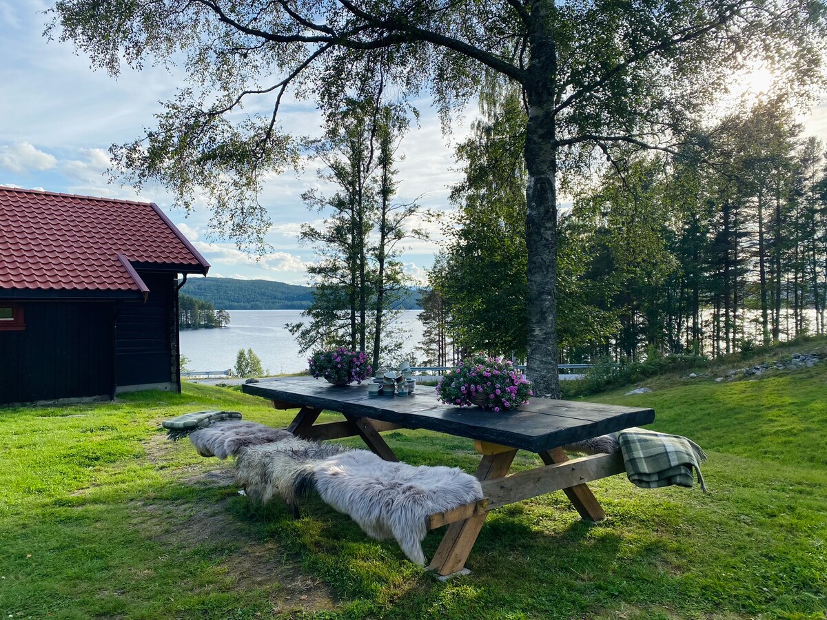 An outdoor seating area features a long wooden table surrounded by benches adorned with soft, light-colored throws. Colorful flower pots sit at the center, with a scenic view of the lake and trees in the background, complemented by the soft afternoon light.