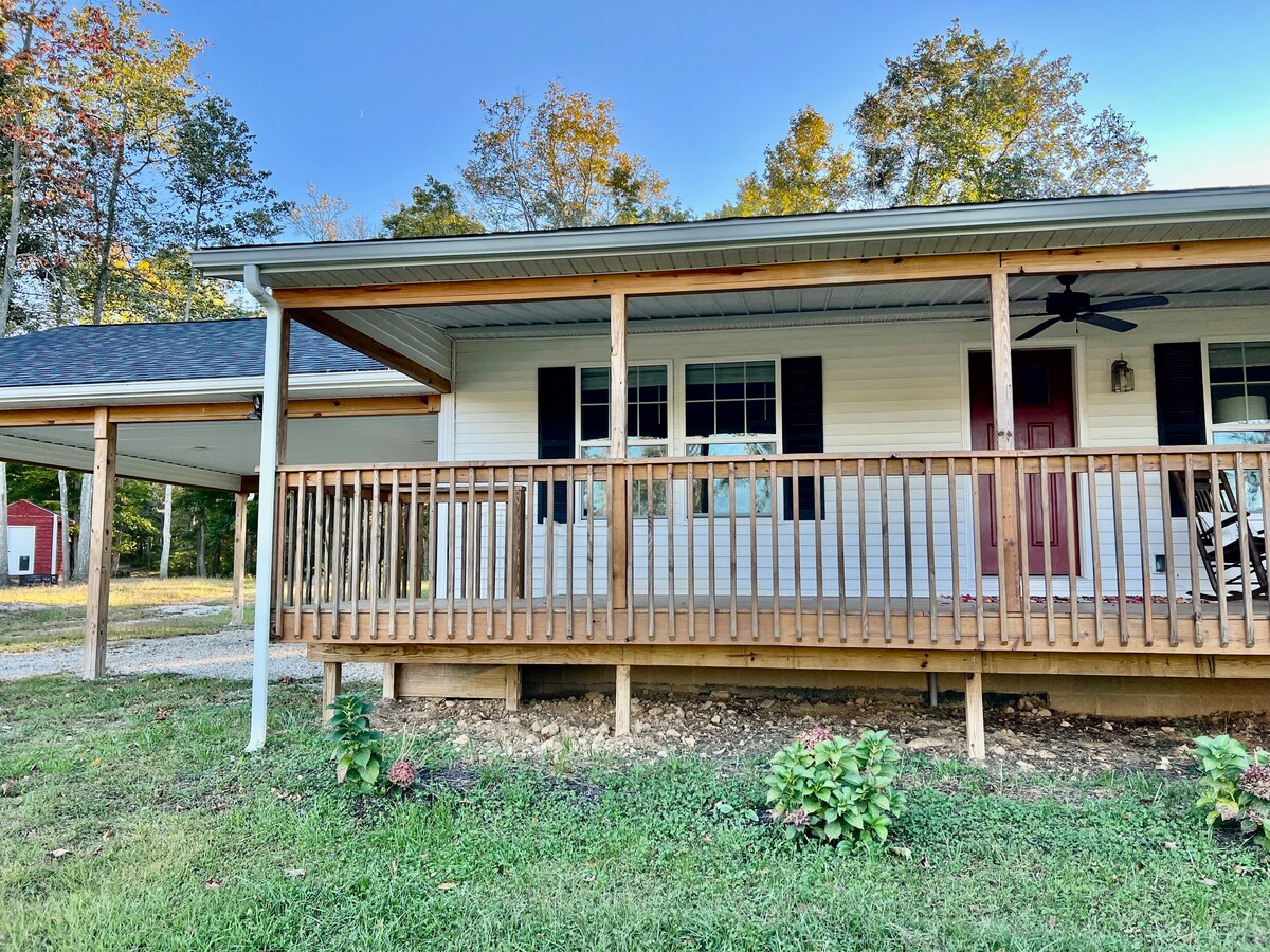 A covered front porch with wooden railings extends along the farmhouse, providing a welcoming entry. The porch features ceiling fans and is flanked by windows, allowing natural light to enter. Green grass and flower beds add to the serene rural setting.