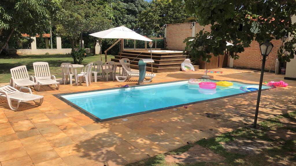 The outdoor area features a rectangular swimming pool surrounded by paving stones. Sun loungers and chairs are arranged nearby, alongside a shaded area with an umbrella. Inflatable floats in various colors can be seen in the pool, and wooden decking is located at the back.