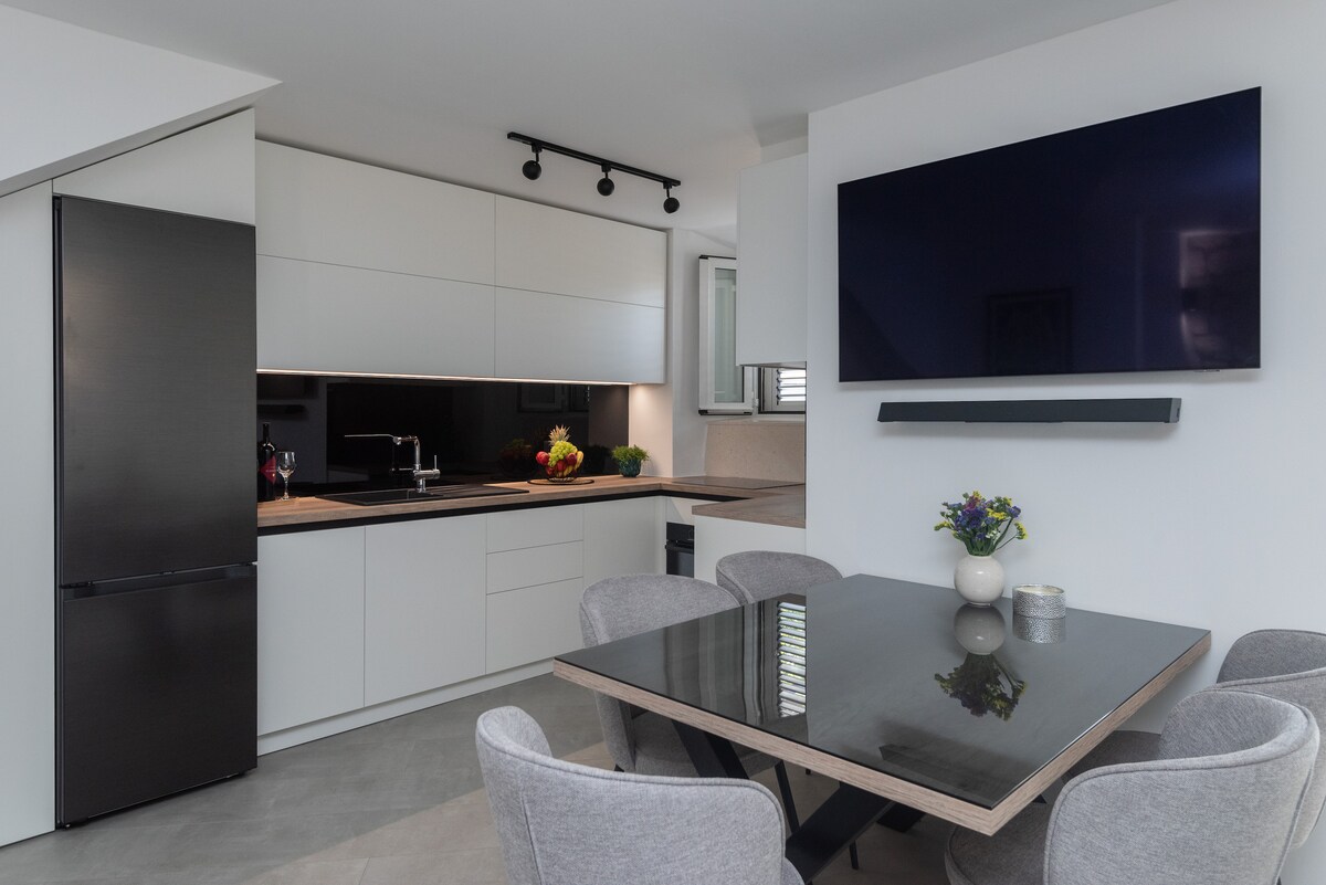A modern kitchen area features sleek cabinetry in neutral tones, complemented by a dark countertop. An elegant dining table with grey chairs is positioned in front of a mounted television. Natural light is allowed in through a window, enhancing the overall bright atmosphere.