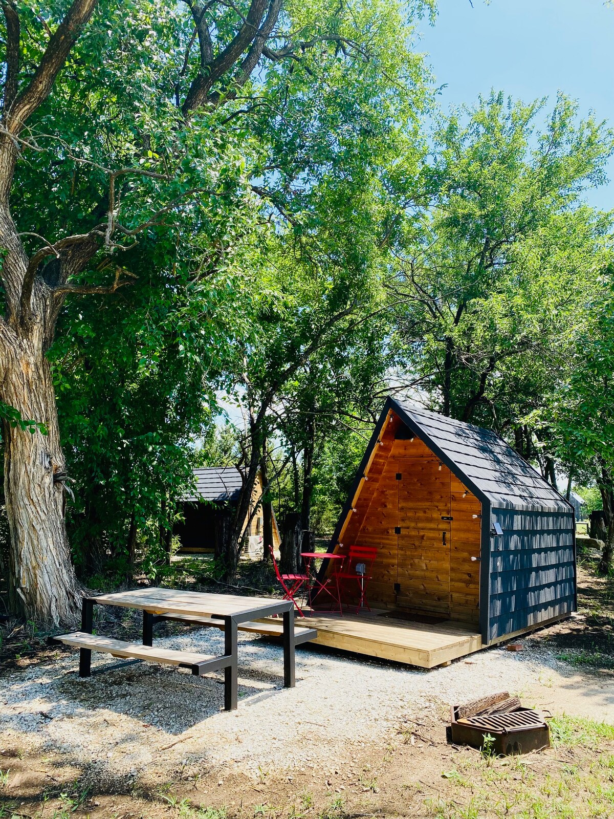A Mini A-Frame cabin is nestled among trees, featuring a wooden porch with a picnic table and two red chairs. A fire pit with grill plate is positioned on the gravel ground nearby. The cabin's sloped roof and natural surroundings create a cozy outdoor atmosphere.