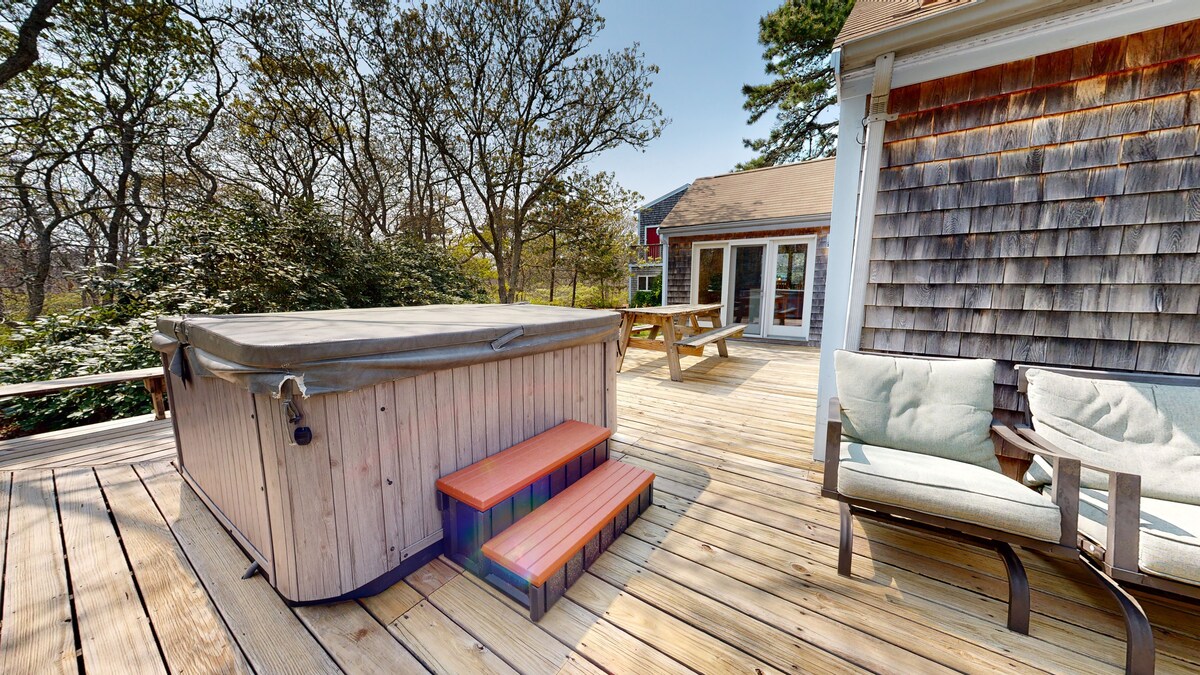 A wooden deck features a hot tub with steps leading up to it. Nearby, a cozy seating area is arranged with light-colored chairs, providing space for relaxation. The surrounding greenery enhances the serene atmosphere of the outdoor retreat.