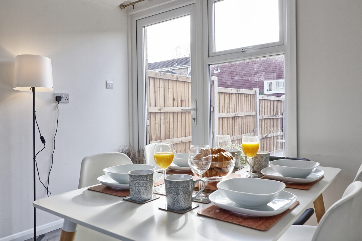 A dining table is set for a meal, featuring plates, bowls, cups, and glasses filled with orange juice. A lamp stands nearby, providing soft lighting. Large windows offer a view of the private fenced garden, bringing in natural light to the space.
