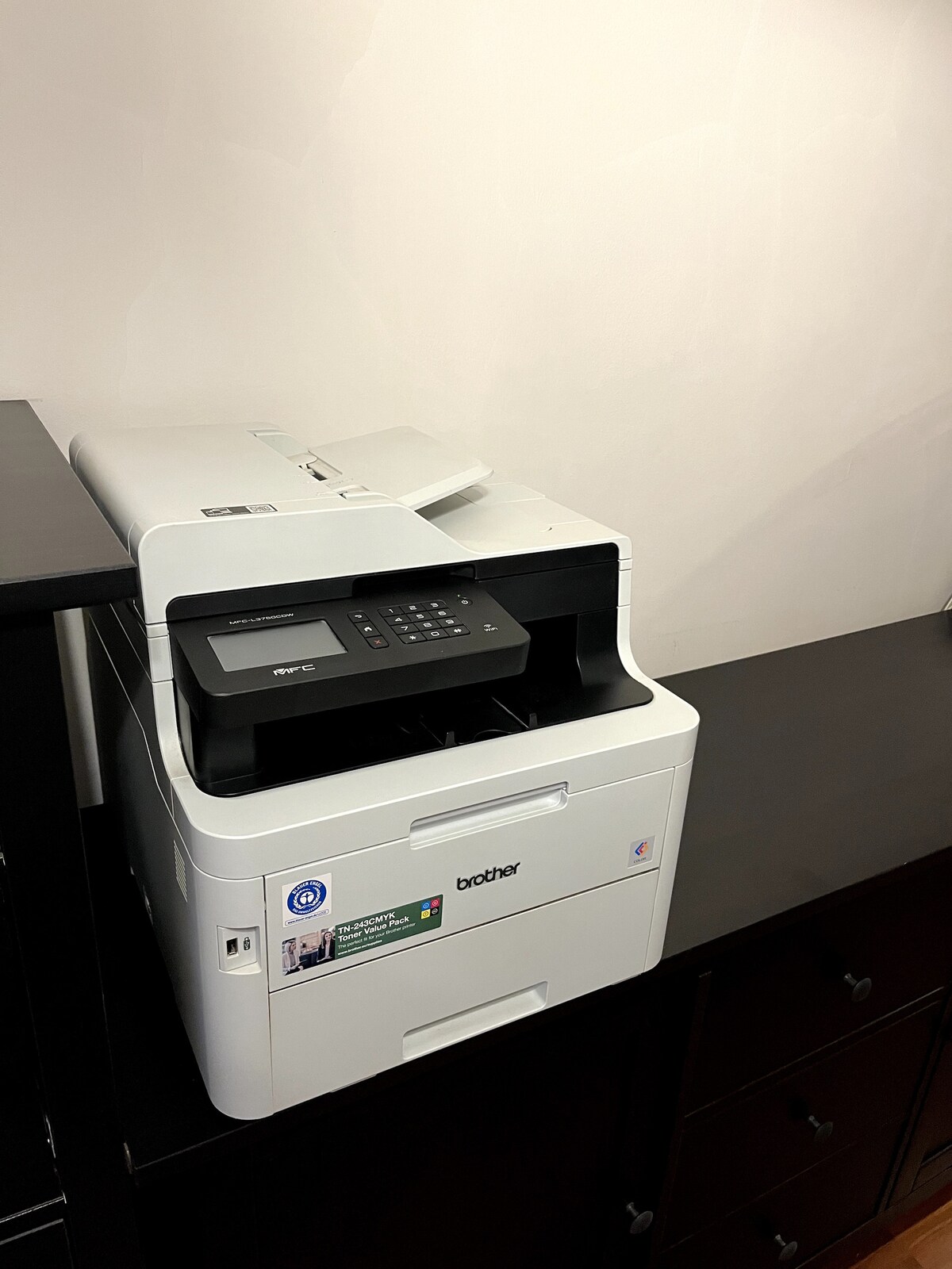 A multi-function printer is situated on a dark wooden surface, featuring a control panel with buttons and a digital display. The printer is designed for various tasks, including printing, scanning, and copying documents.