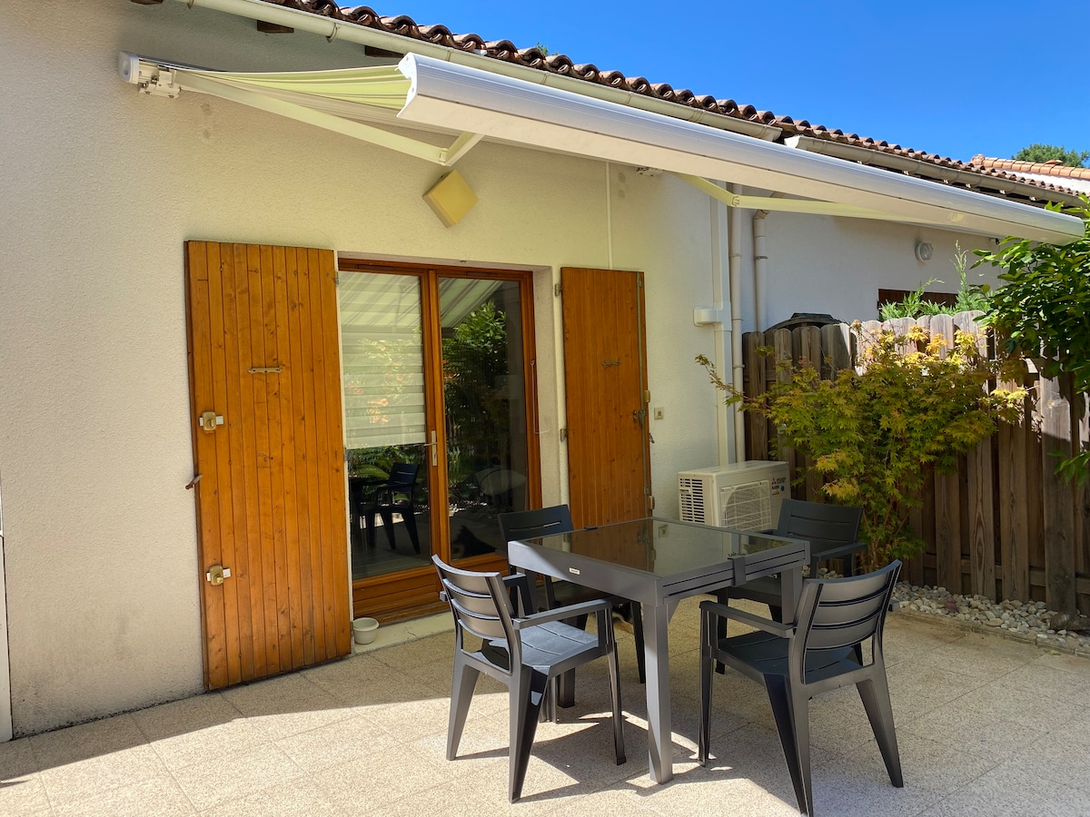 An outdoor dining area features a dark table surrounded by several matching chairs. Light reflects off a surface in front of large glass doors that open to an interior space. Brown wooden shutters are positioned on either side of the doors, with greenery visible beyond.