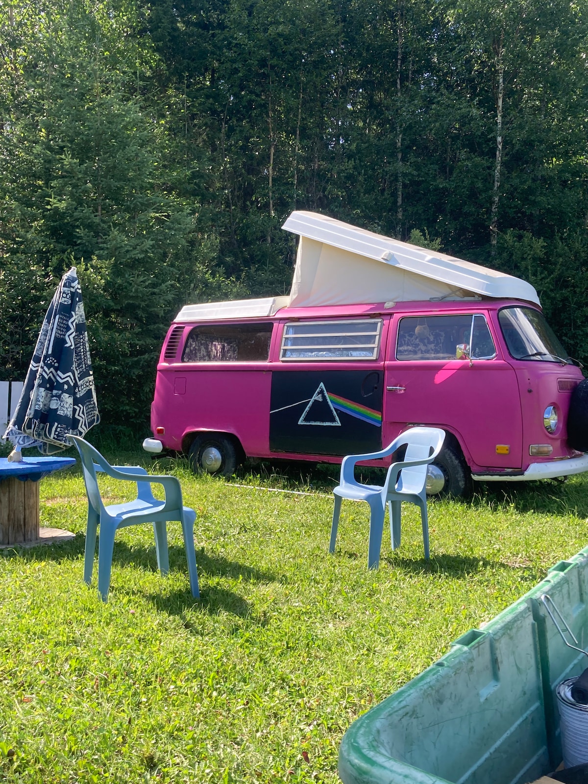 A vintage pink camper bus is parked on a grassy area, featuring a raised pop-top. Two light blue chairs are positioned nearby, while a patterned umbrella adds shade. A wooden table is visible in the foreground, accompanied by a green bin on one side.