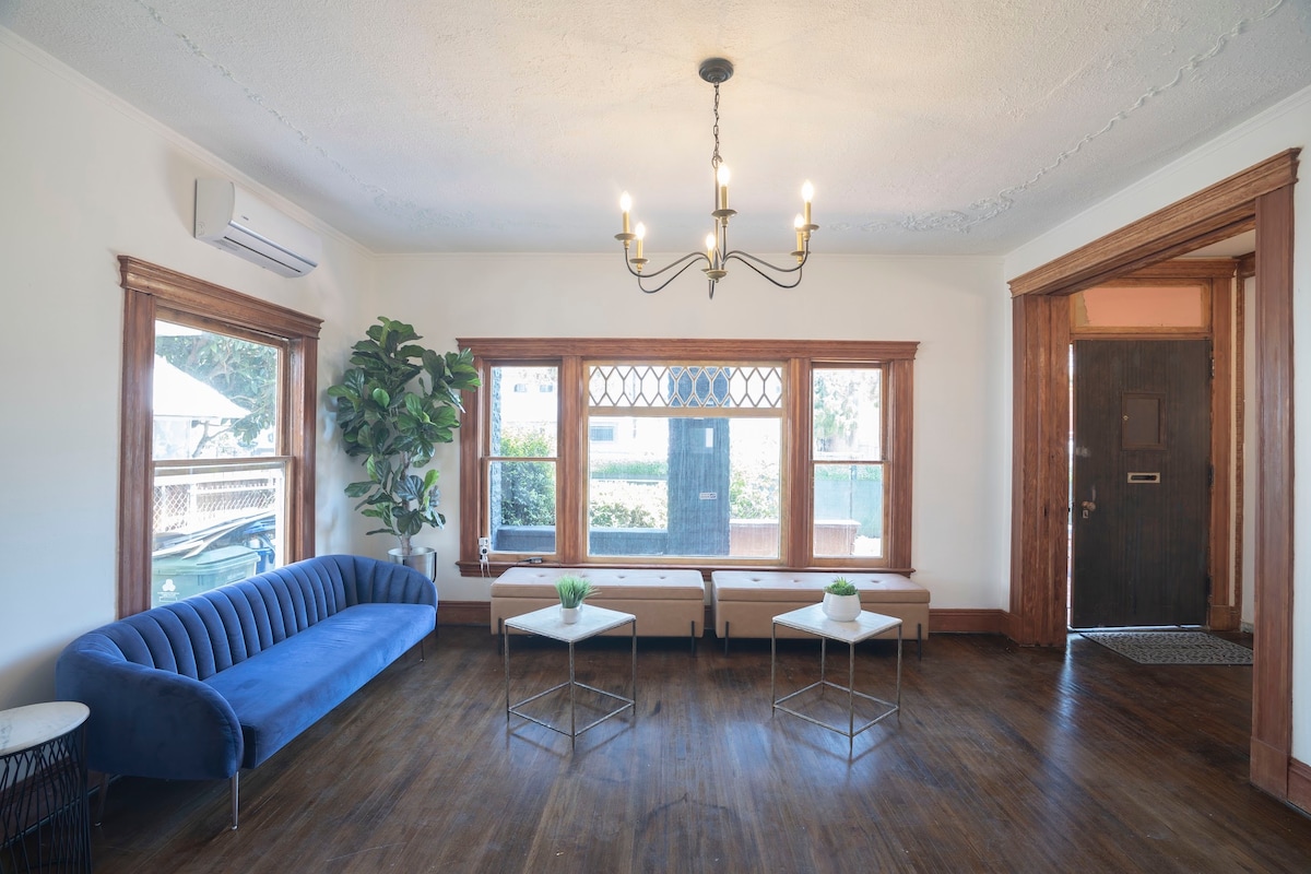 A spacious common area is illuminated by natural light from large windows. A deep blue velvet sofa is positioned alongside two small glass tables. The hardwood floor contributes warmth, while decorative plants add a touch of greenery. A chandelier provides overhead lighting.