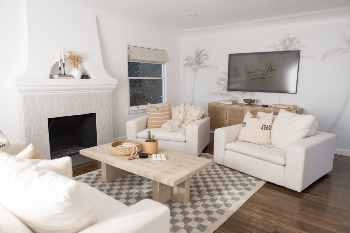 A bright living room features a cozy arrangement of white sofas surrounding a light wooden coffee table. A patterned rug lies beneath. A flat-screen television is mounted on the wall, and natural light enters through window treatments, enhancing the neutral color palette.