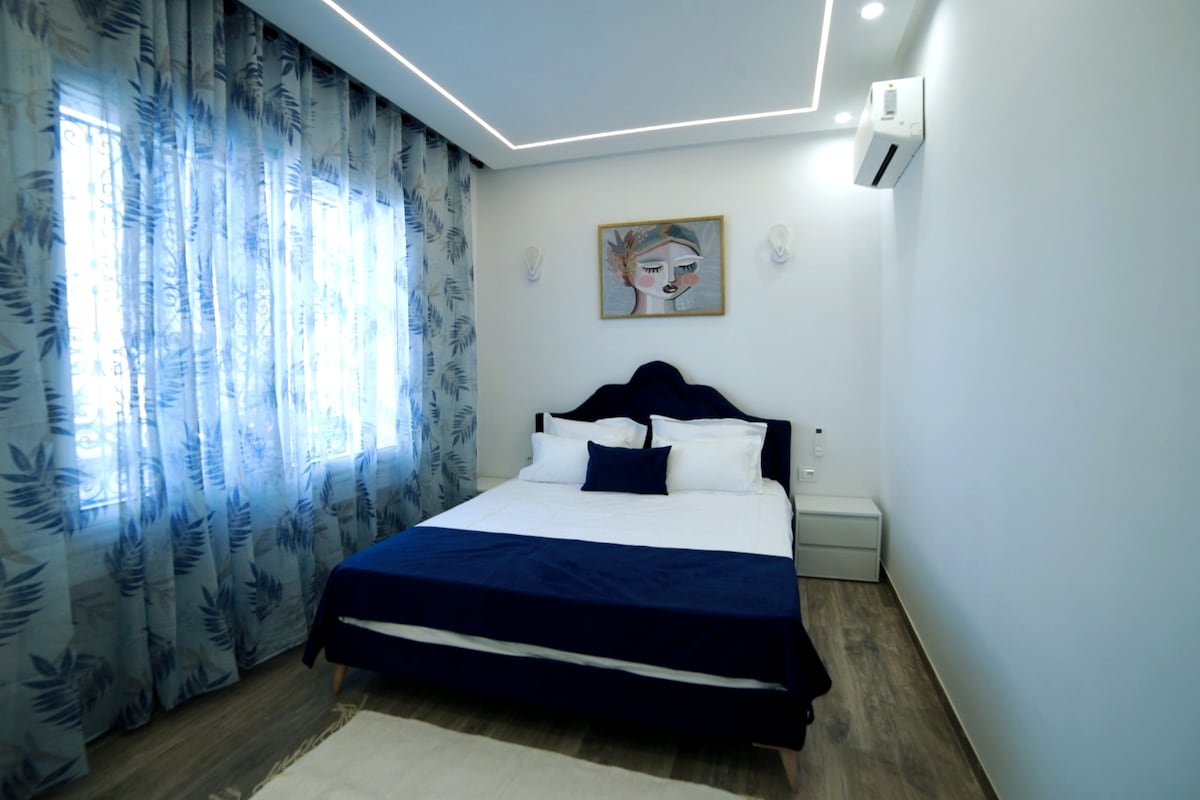 A bright bedroom features a spacious bed with crisp white linens and a navy blue throw pillow. Sheer, patterned curtains frame the window, allowing natural light to illuminate the room. An air conditioning unit and wall art can be seen, enhancing the modern design.
