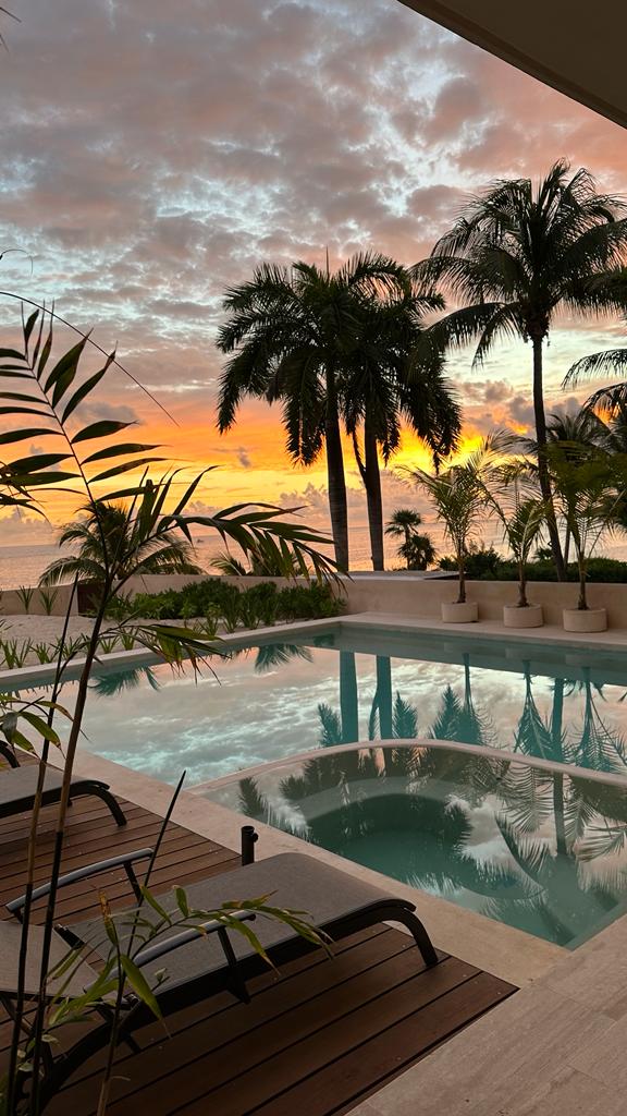 A serene view is captured from the villa, showcasing a tranquil pool reflecting the vibrant colors of a sunset. Silhouettes of palm trees frame the scene, enhancing the ambiance of relaxation by the beachfront.