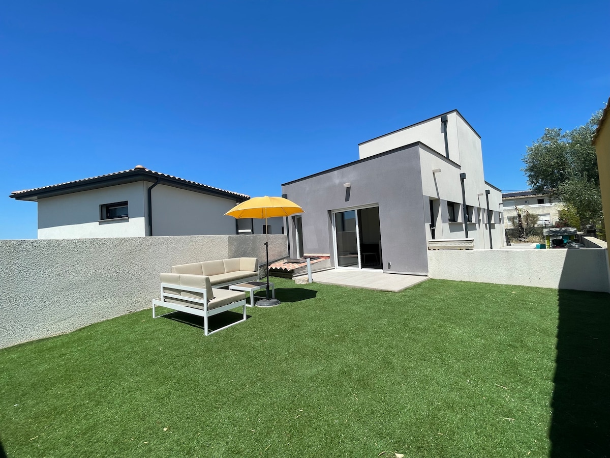 A private garden area is featured, presenting a green lawn and a shaded seating arrangement under a yellow umbrella. The modern building is visible in the background, showcasing large glass doors that lead outdoors, enhancing the connection to the sunny environment.