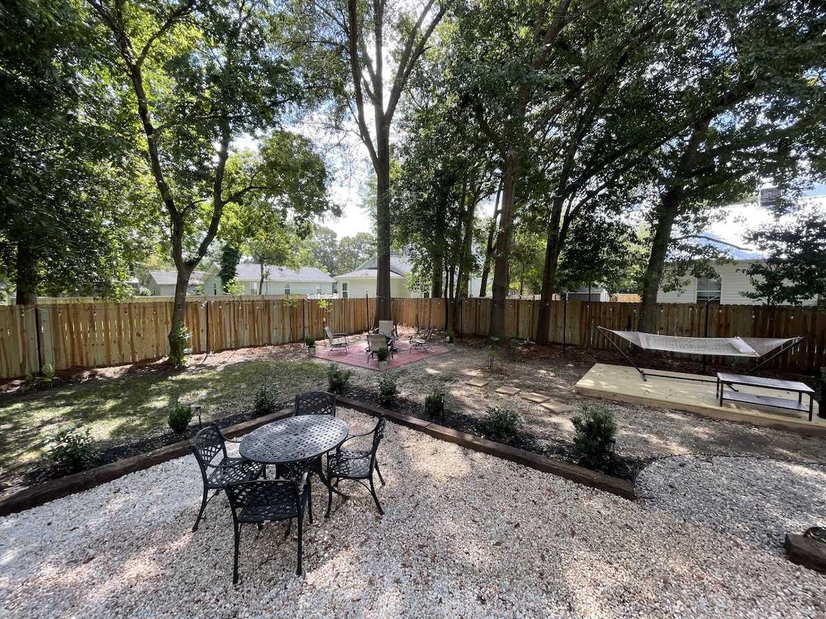 The spacious backyard features a gravel area with a round table and four chairs, surrounded by landscaped borders. A hammock is positioned under the trees, with additional seating areas visible. Tall trees provide shade and privacy, creating a serene outdoor environment.