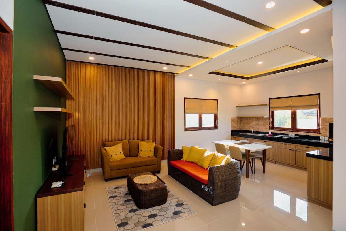 A spacious living area is presented with a comfortable seating arrangement featuring a yellow sofa and a vibrant orange accent chair. A dining table is visible, along with modern cabinetry in the kitchen. Natural light enters through two windows adorned with shades.