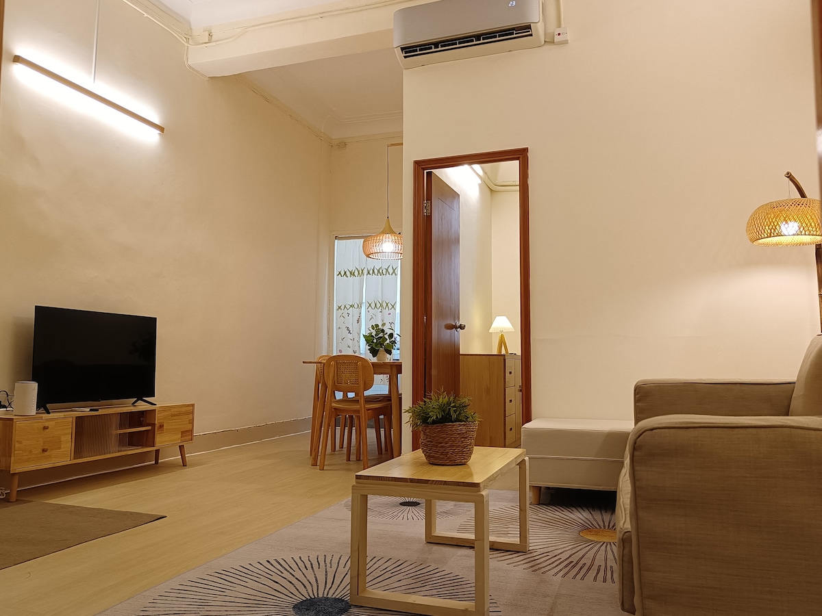 The living and dining area features a light and airy space with high ceiling. A comfortable beige sofa is positioned next to a small table and potted plant. A wooden dining table with chairs is visible in the background, along with a television on a sleek stand.
