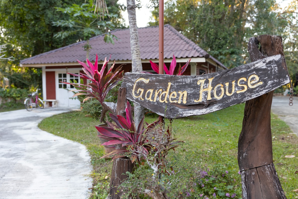 A wooden sign marked 'Garden House' is prominently displayed, flanked by lush tropical plants with vibrant pink leaves. The eco garden house is visible in the background, featuring a sloped roof and large windows, surrounded by greenery.
