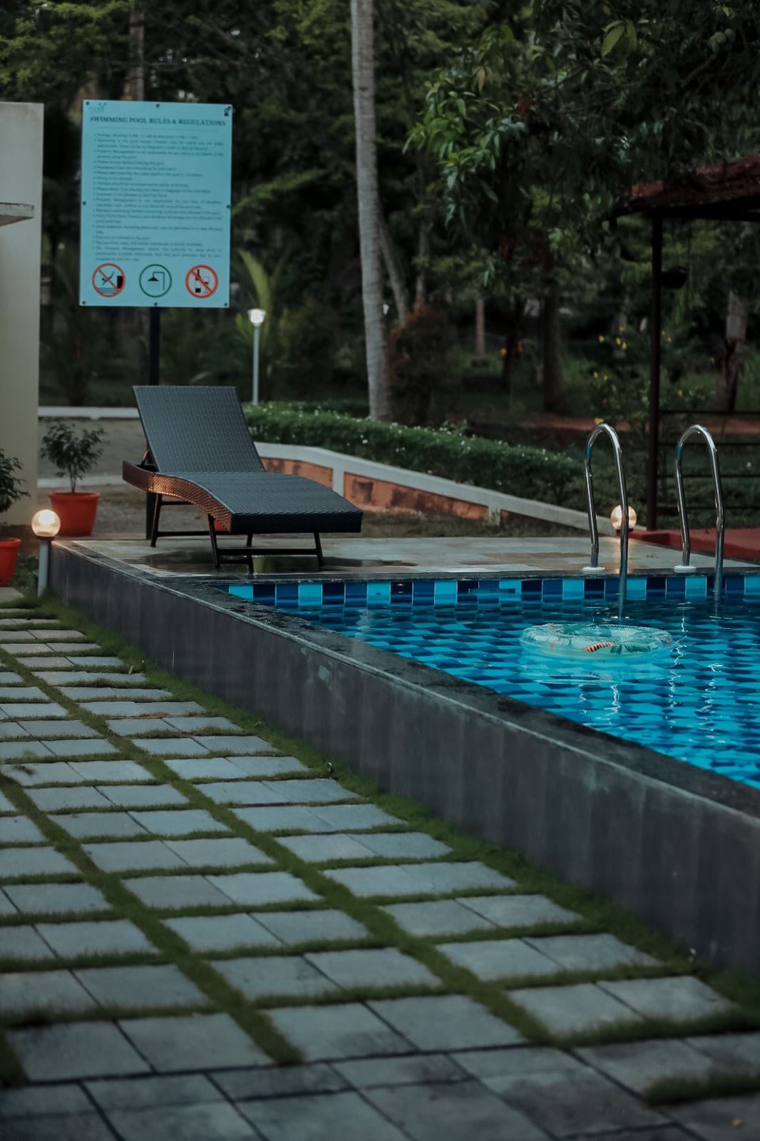 A well-maintained pool area is featured, highlighted by a lounge chair positioned beside the water. The pool is adorned with vibrant blue tiles. Lush greenery and decorative lights surround the space, providing a serene ambiance.