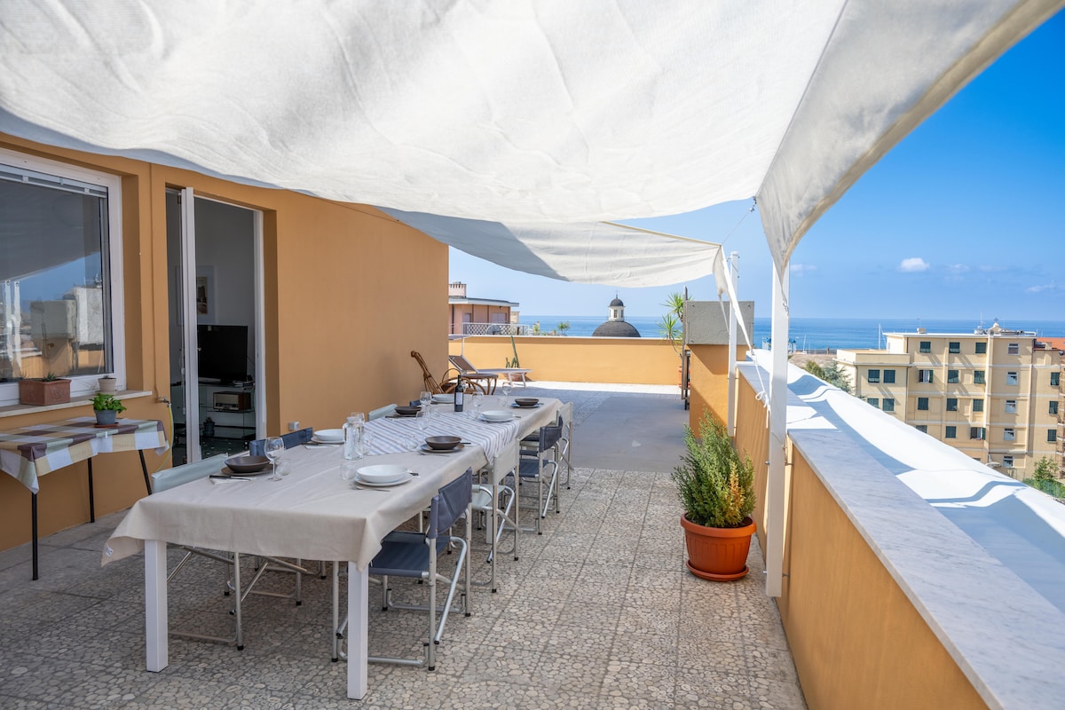 An expansive terrace offers a sea view, featuring a large dining table set for outdoor meals. Sunlight filters through a canopy, casting soft shadows on the tiled floor. A potted plant adds a touch of greenery, while the nearby building outlines the vibrant coastal setting.