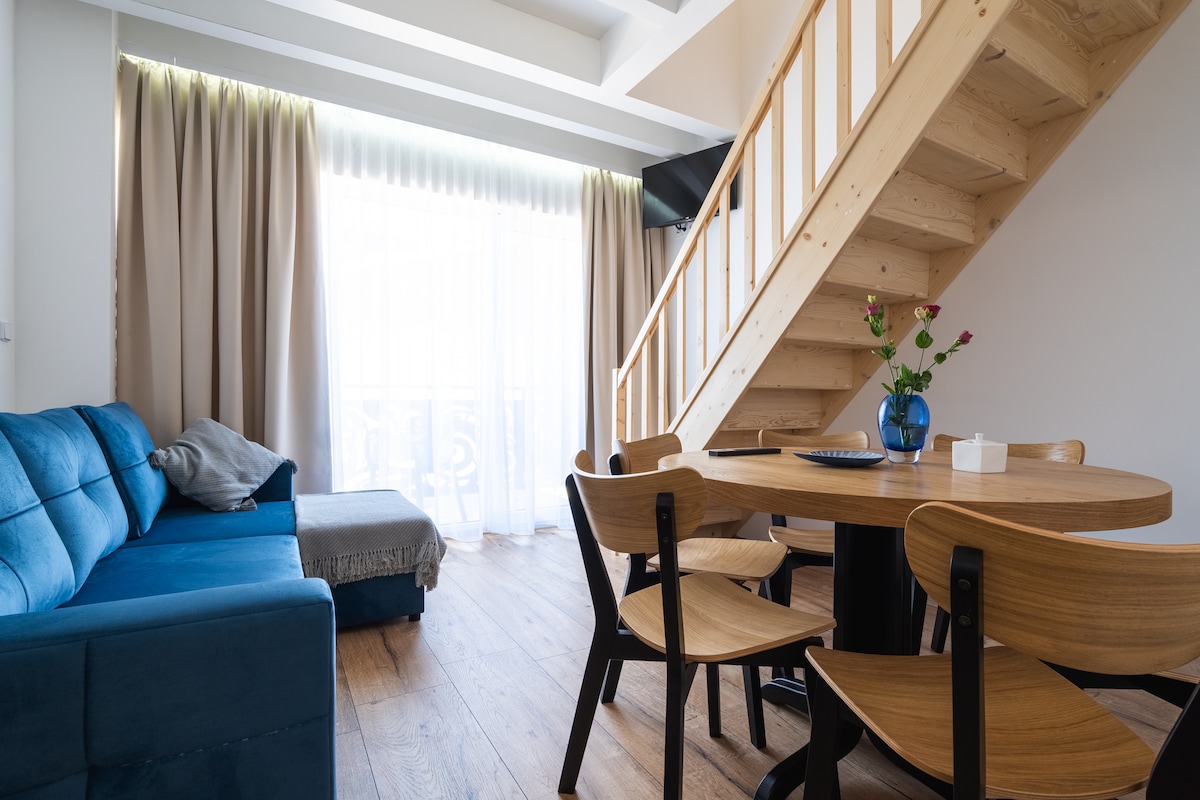 A spacious living area is featured, showcasing a blue sofa with throw pillows and a small side table. A wooden dining table is surrounded by four chairs. Natural light enters through large curtains, while a staircase leads to an upper level.