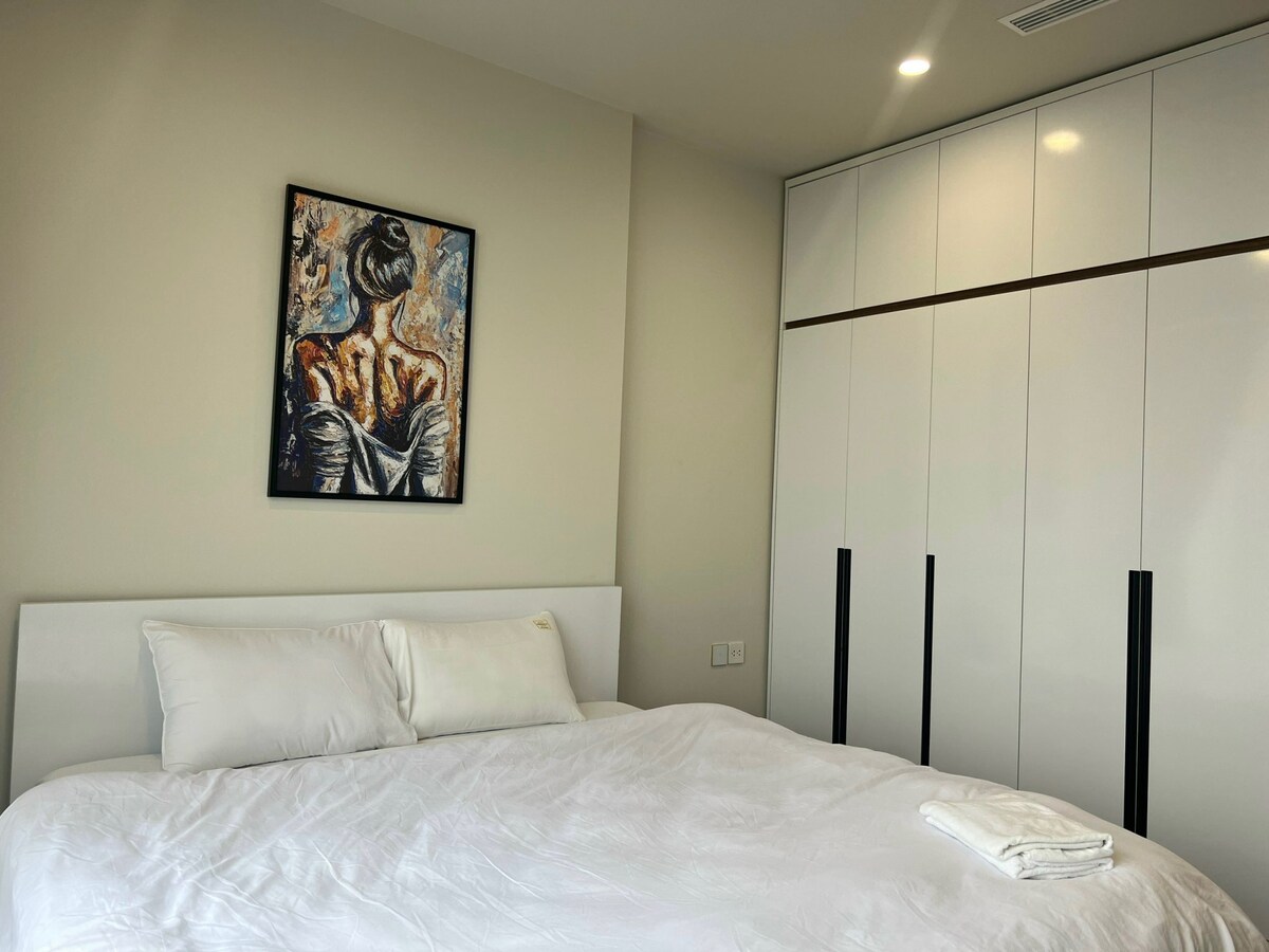 A spacious sleeping area features a large bed dressed in white linens. A striking piece of wall art depicts a woman in a stylish pose. A sleek, white wardrobe with black handles stands against the wall, providing ample storage.