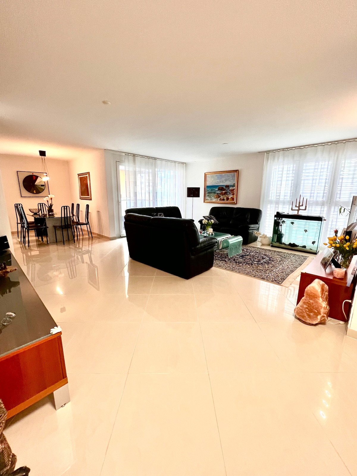 A spacious living area features modern furnishings, including two black leather sofas facing a decorative coffee table. Natural light filters through sheer curtains. The open layout connects to a dining area with a glass table and chairs, enhancing the room's inviting ambiance.