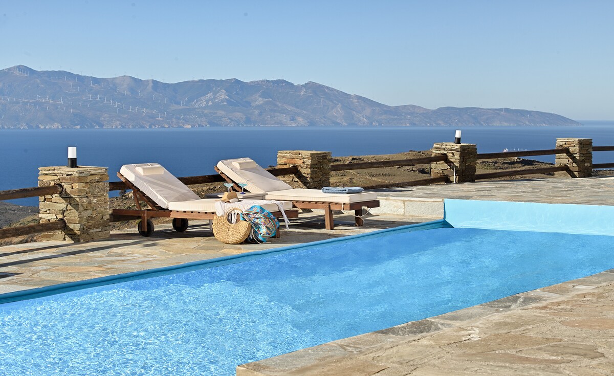 A serene pool area features inviting blue water reflecting the sky. Two lounge chairs are positioned nearby on a stone deck, accompanied by a wicker basket containing beach accessories. The expansive view showcases the sea and distant mountains, enhancing the tranquil atmosphere.