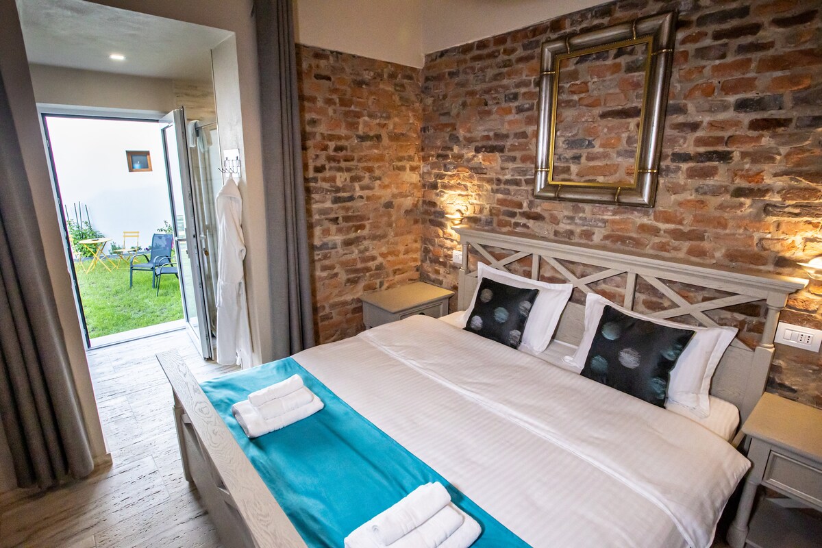 A stylish bedroom features a spacious bed with a turquoise throw and neatly arranged white towels. Exposed brick walls add character. A door provides direct access to the shared garden, allowing natural light to fill the room, complemented by bedside lamps.